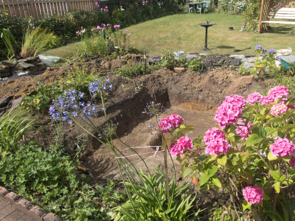 Gartenteich anlegen – Ausgehobene Grube für einen Teich, umgeben von Blumen und Gewächsen.